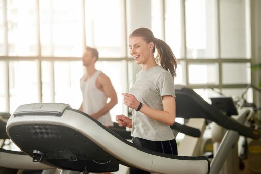 Person with ponytail in grey t-shirt runs on treadmill in gym, wearing headphones